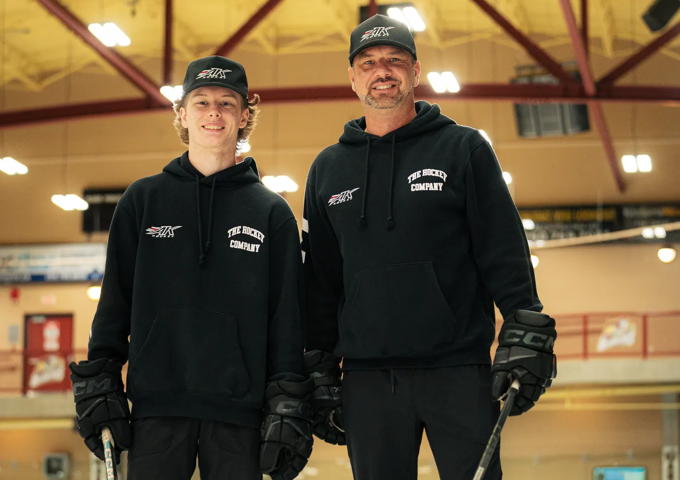 Two AK Hockey coaches in AK Hockey hats and hoodies standing on the ice